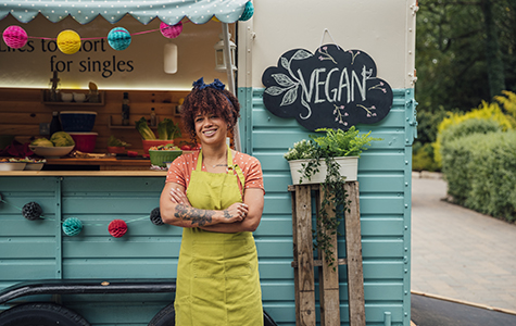 Proud small business owner stands in front of her vegan food cart with her arms folded.