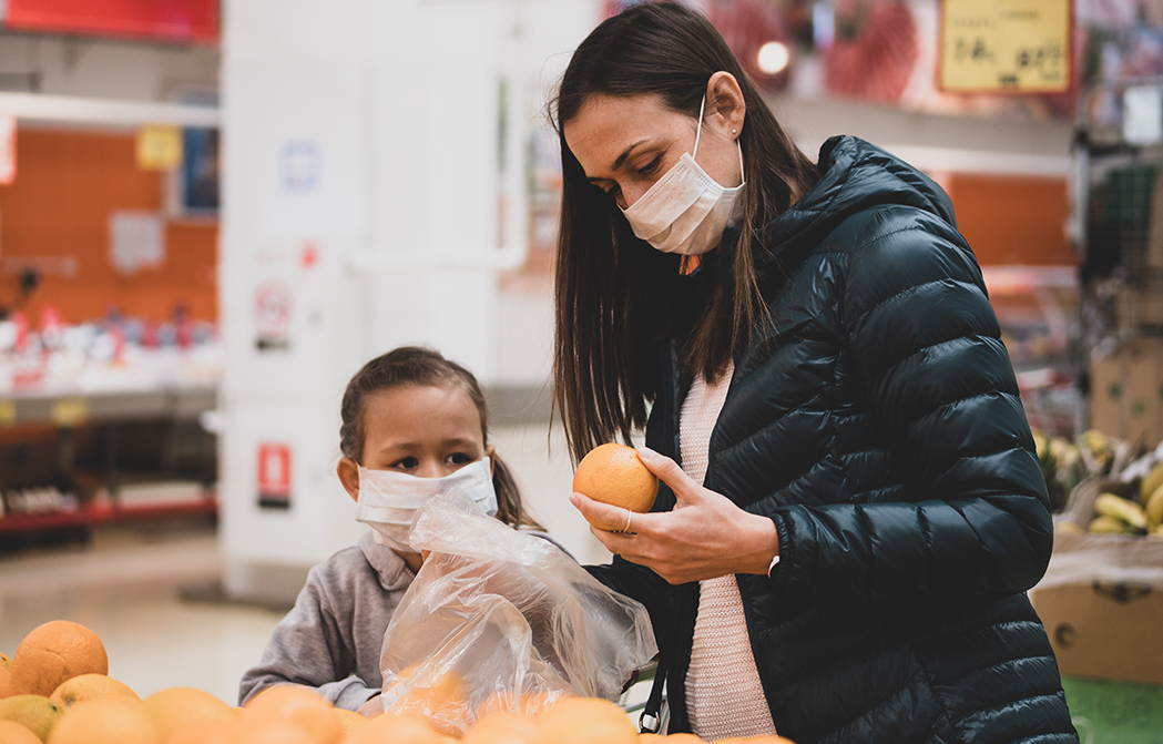 Young woman with child girl in medical masks buys a oranges at supermarket