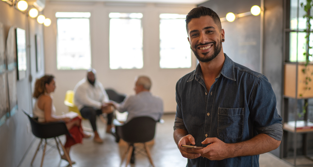 How Small Business Owners Can Teach Their Employees About Cybersecurity_Young Man holding cell phone with coworkers in background