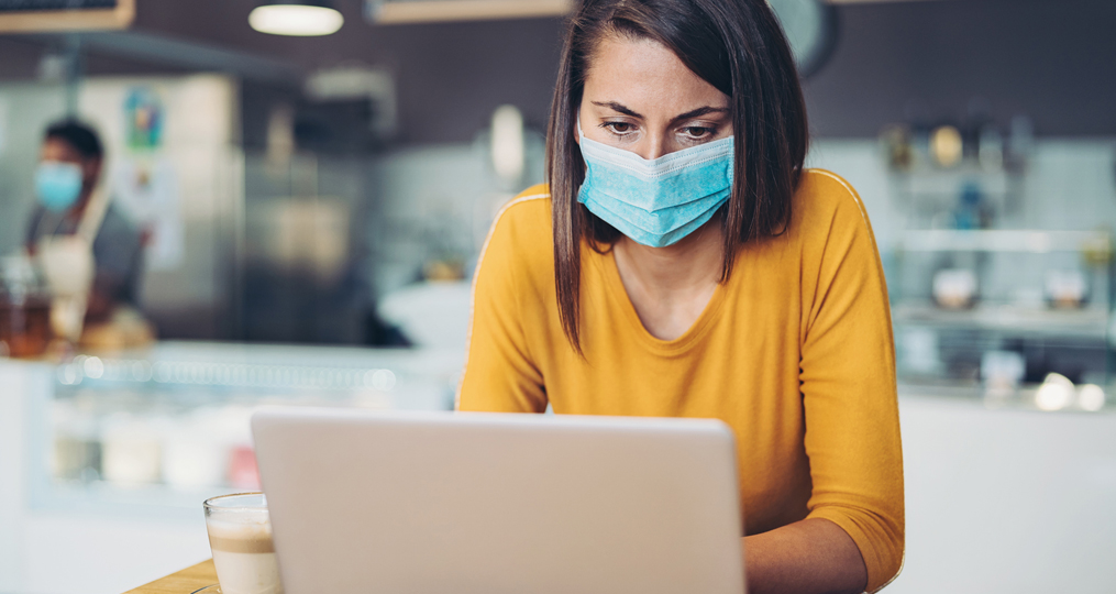 Protect Yourself from COVID-19 Fraud and Scams_Professional Woman working on her computer at a local coffee shop wearing a mask