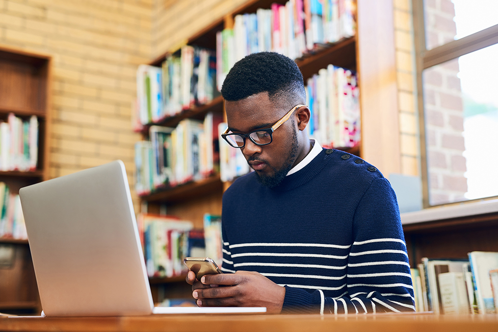 Young man at school texting while working on a laptop