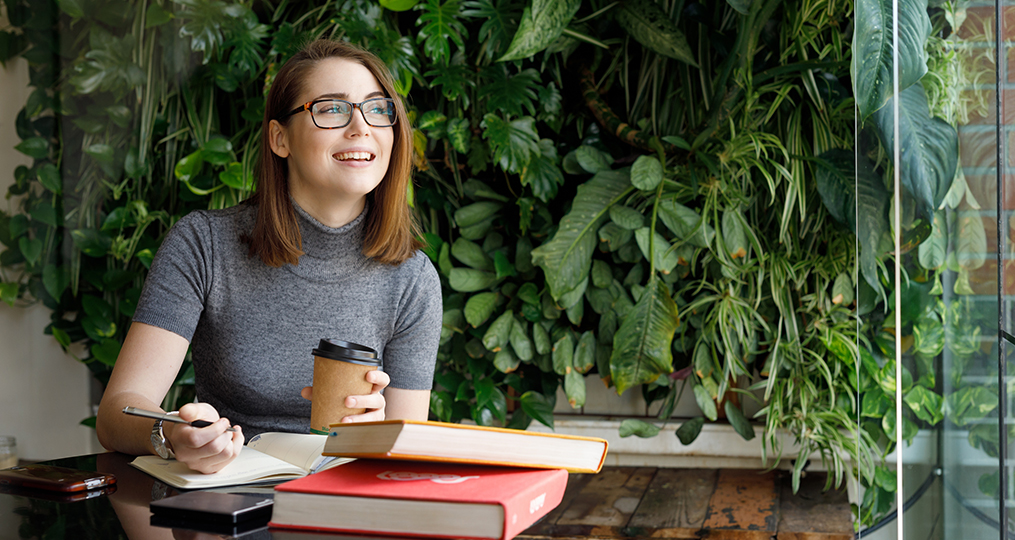 4 financial habits to develop in your 20s - young woman sitting at a desk taking notes and looking out the window