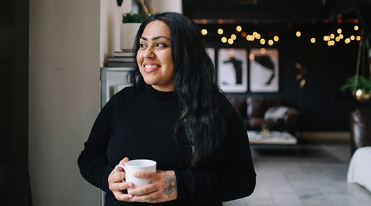 Commit to Getting Financially Fit in 2021_young woman standing in her apartment with a cup of coffee thinking about the future