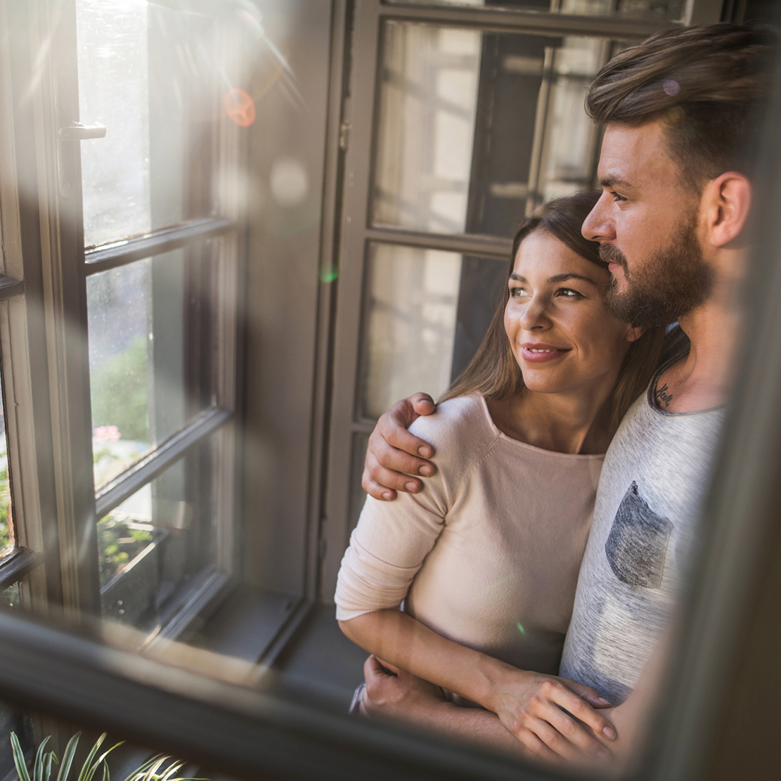 couple standing at the window of their new home looking out into their front lawn