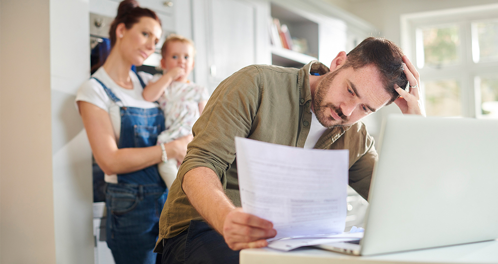 managing personal finances during a crisis_Man sitting at kitchen table reviewing bills while family looks on