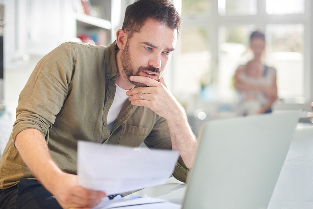 managing personal finances during a crisis_Man sitting at kitchen table reviewing bills making a plan while family stands in background