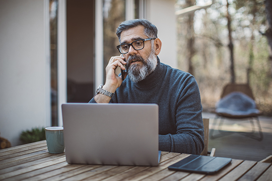 mature man on cell phone at laptop on patio and looking concerned
