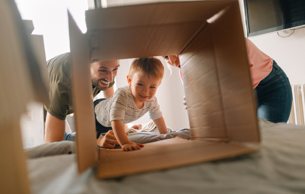 young couple watching their son crawl through a moving box on their bed