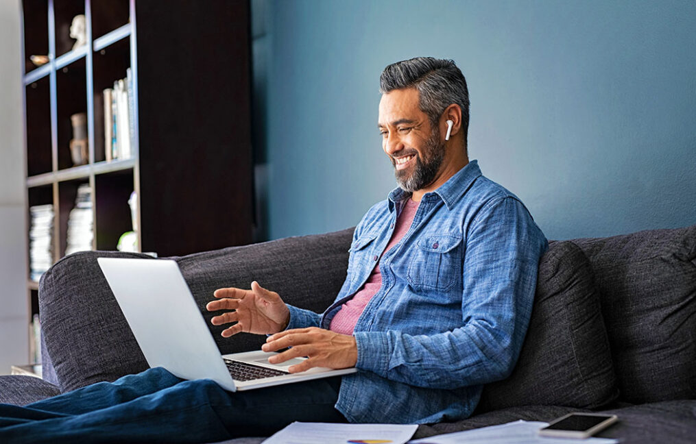 Happy mature indian man sitting on couch while doing a video call with laptop. Mixed race businessman sitting on sofa and making a video conference with wireless earphones while working from home, new normal concept. Smiling mid adult ethnic business man doing online video with his colleagues.