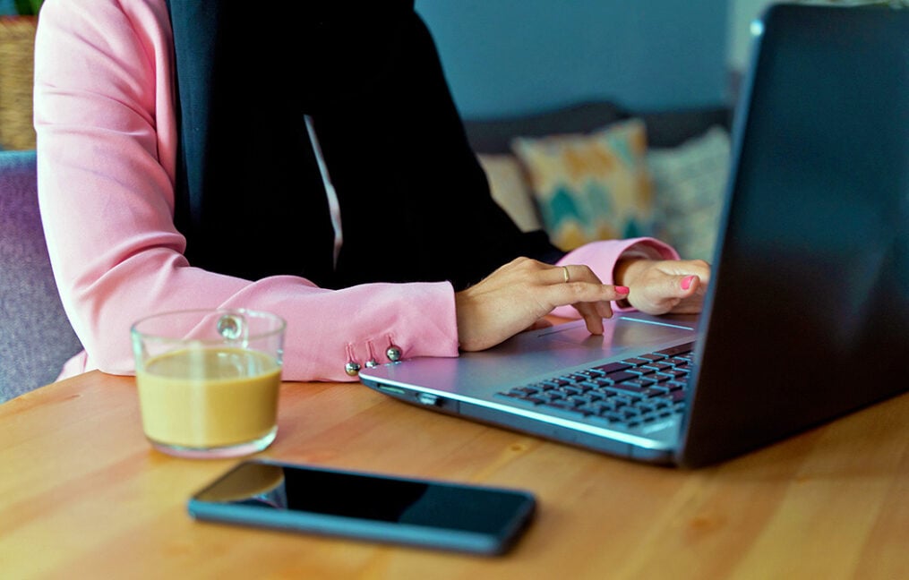 Close up of woman's hands typing on laptop keyboard with phone and coffee cup on table in foreground