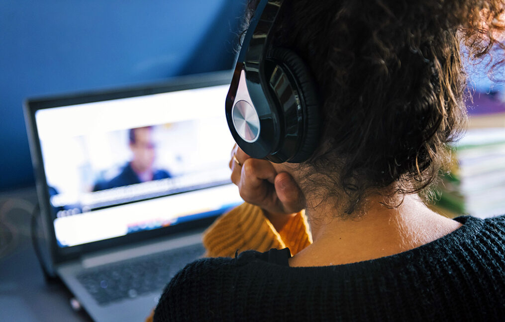 Gen Z woman on over-ear headphones looks at computer screen with video conference image