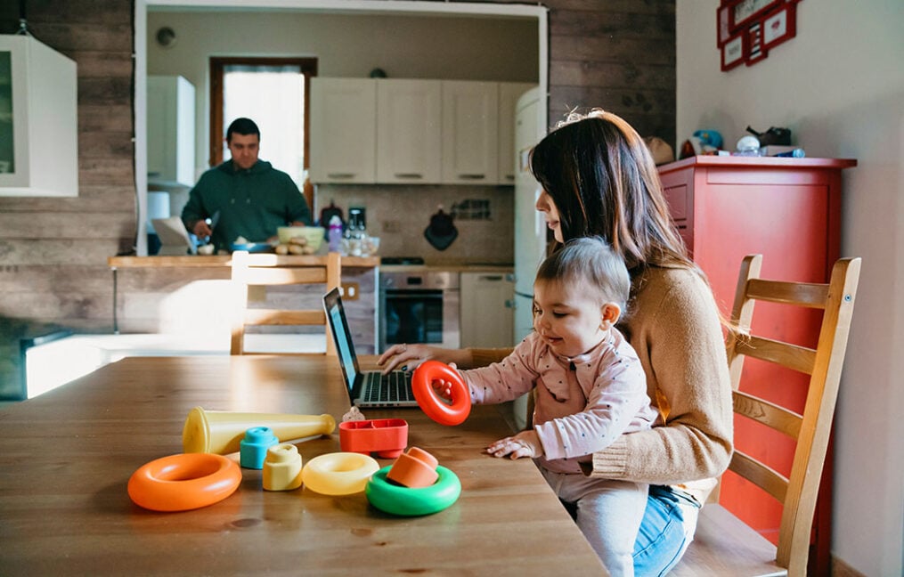 Woman with baby on lap at dining table and reviewing laptop with husband in the kitchen in the background