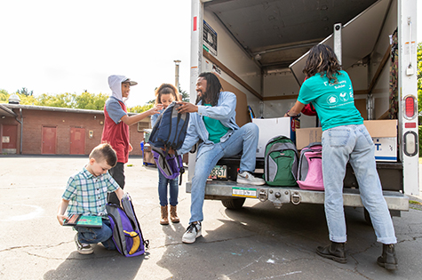 Man volunteering hands out backpacks and school supplies to young children.