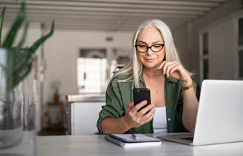 Woman at desk with laptop, looking at phone and finances.