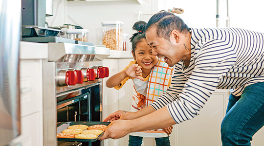 asian man pulling cookies out of oven while daughter joyfully watches