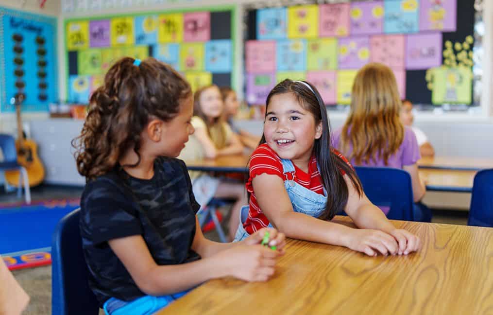 two little young girls sitting at classroom table talking smiling onpoint prize 2025