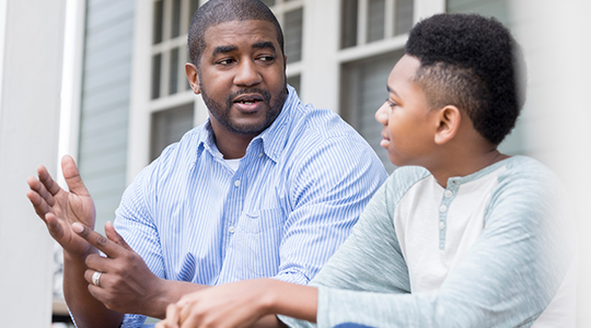 Caring mid African American father gestures as he gives advice to his preteen son. They are looking at one another. They are sitting on the front porch of their home.