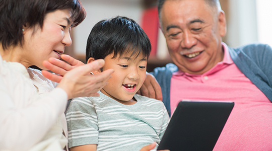 Young boy using his digital tablet whilst his grandparents watch and offer encouragement.