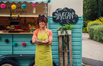 Outside a food truck where a mixed race female hipster stands with her arms folded smiling to the camera.