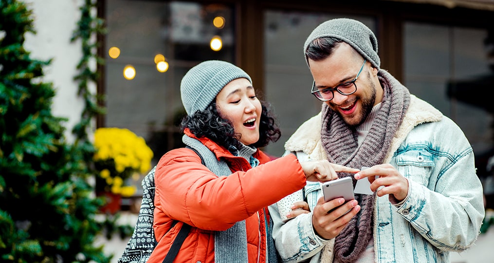 avoid overspending this holiday season-couple standing outside looking at mobile phone during the holidays