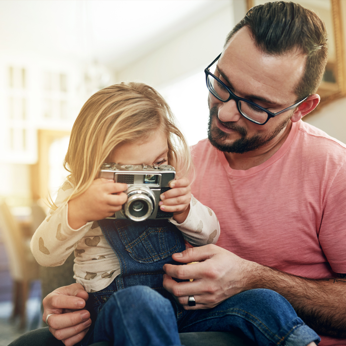 National Credit Union Youth Month_daughter sitting on fathers lap holding a camera and taking a picture