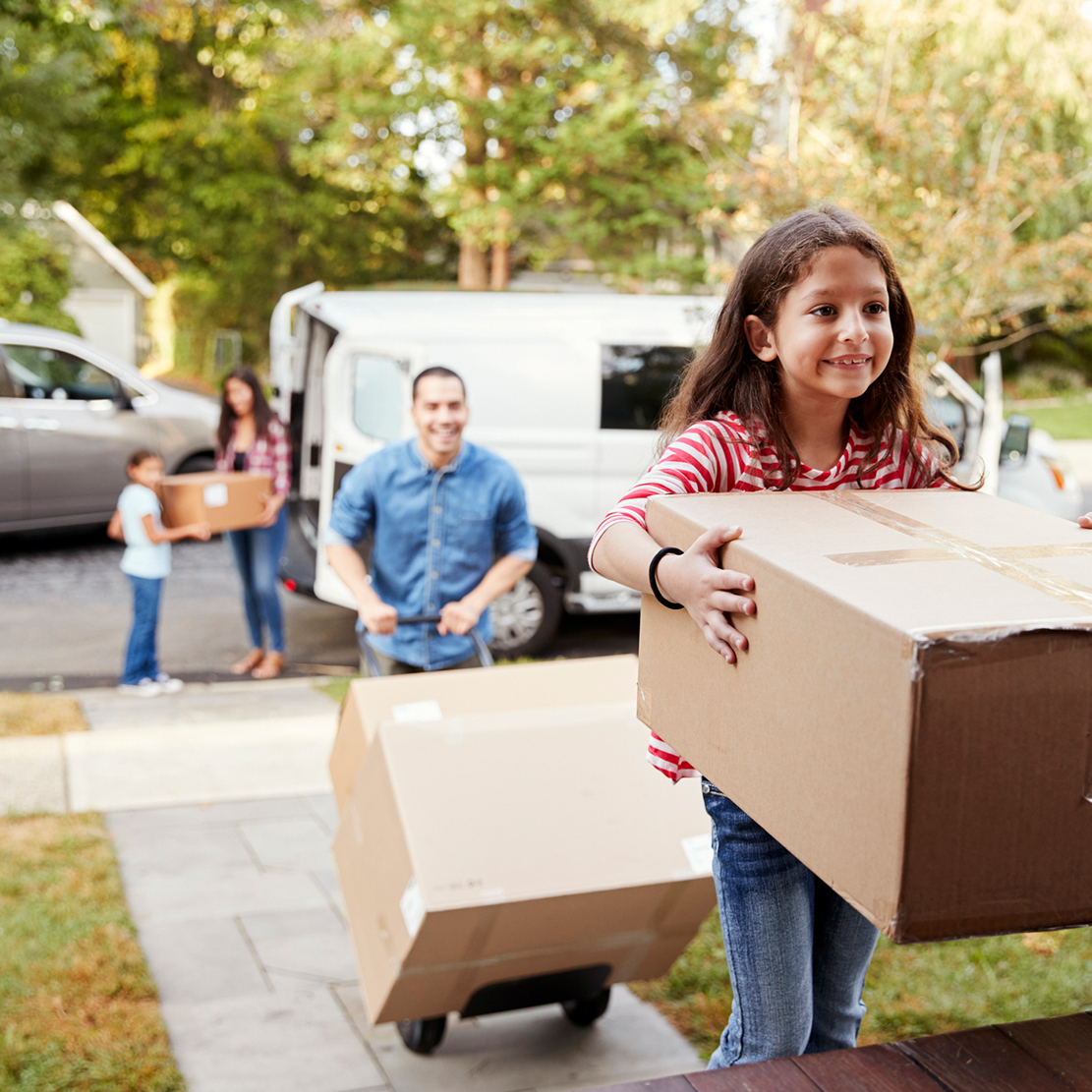 young girl carrying a box into her new home - with the rest of family unloading boxes in the background
