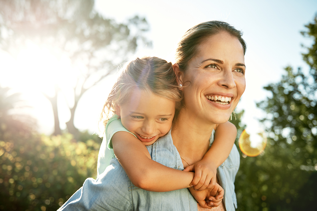 mom giving her young daughter a piggy back ride while they both smile and laugh