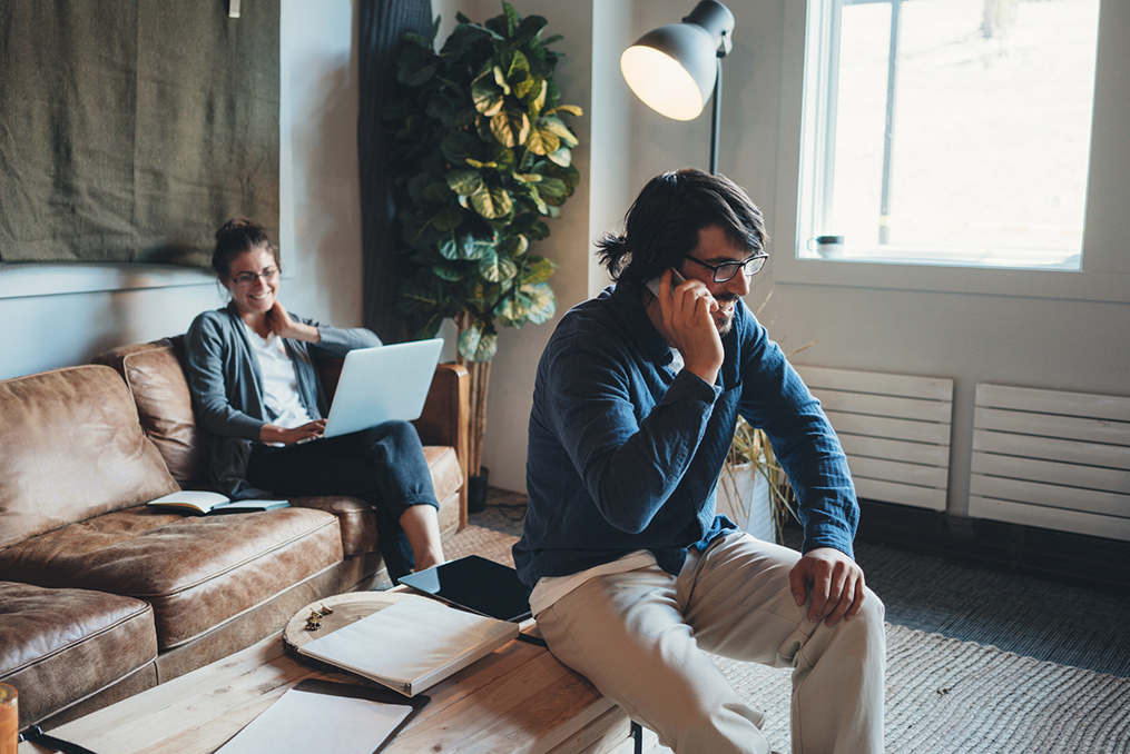 woman sitting on her couch with her laptop while man sits on coffee table and makes a phone call