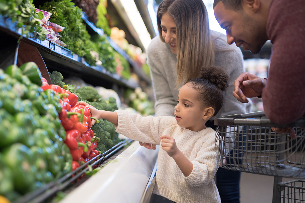 young child helping her parents pick out vegetables at the grocery store