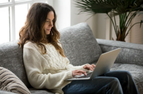 Smiling young woman sitting on sofa using laptop communicating online